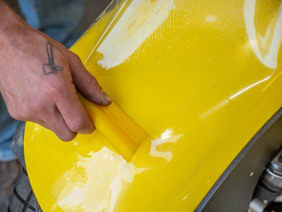 A squeegee smoothing clear protective film over a yellow fender.