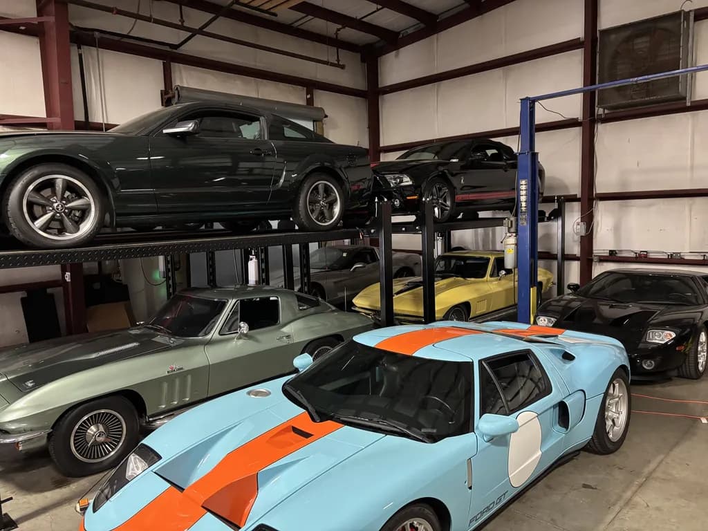 A Ford GT in blue and orange livery parked beside a silver Corvette Sting Ray, with more cars racked above.