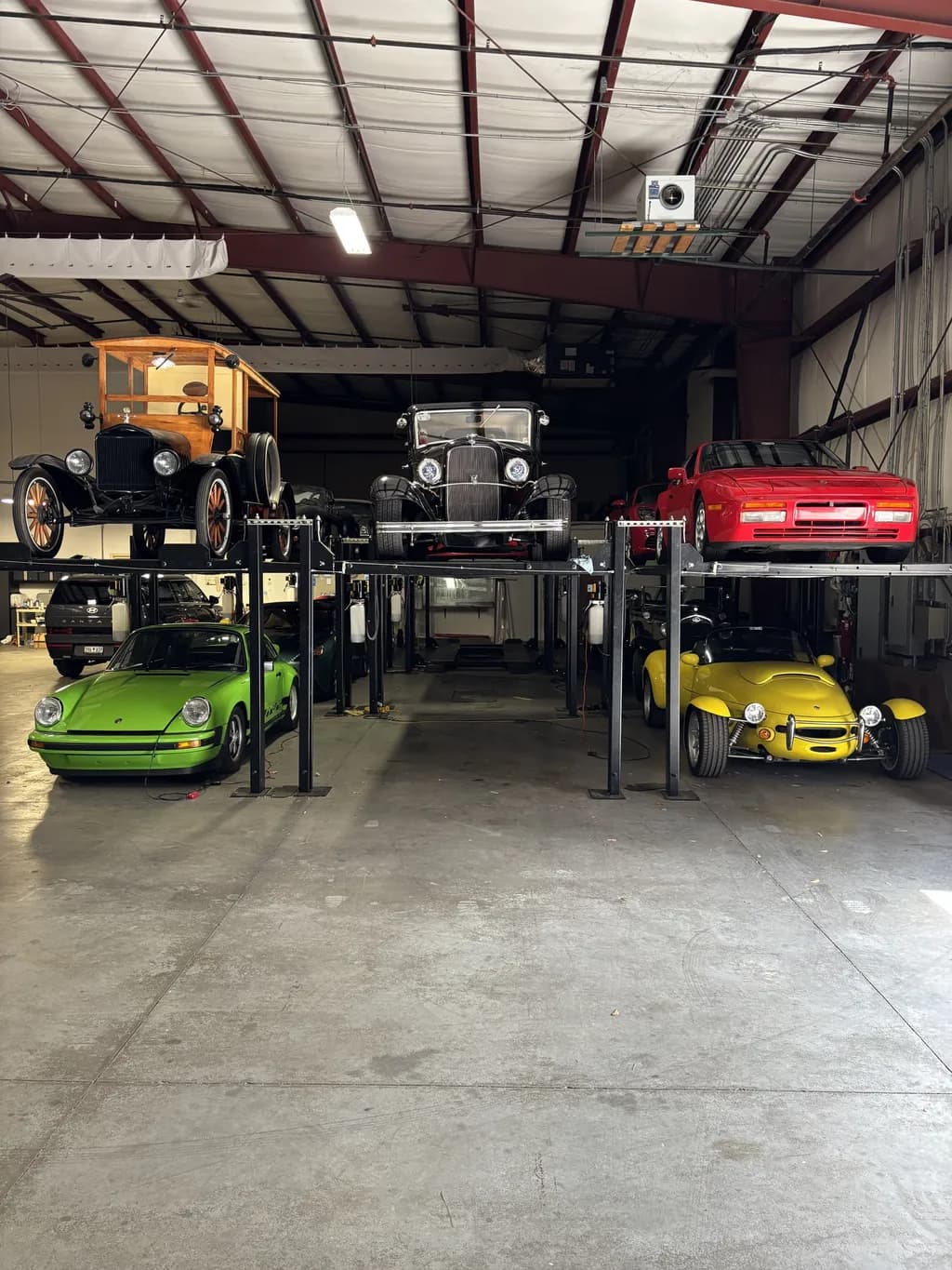 Vintage cars on racks above a green Porsche 911 and a yellow roadster in the storage building.